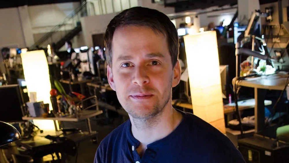 A man with short hair, the start of a beard, and wearing a blue button down shirt looking at the camera.