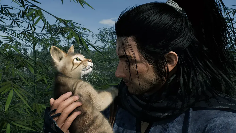 A person with long black hair is holding a small, light brown cat. The cat is looking upwards with its mouth slightly open, showing its teeth. The background consists of green bamboo plants and a clear blue sky.