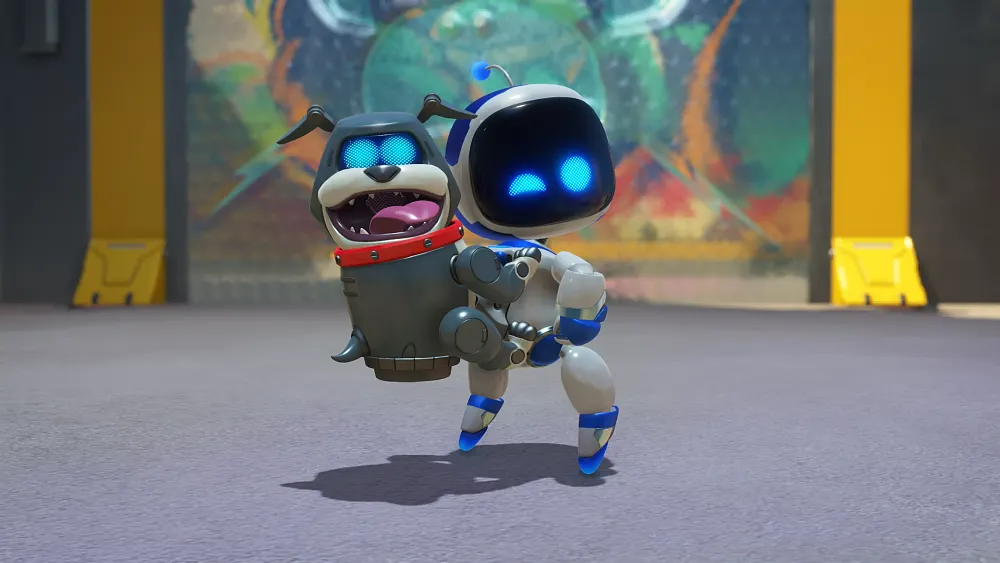 A small, white and blue robot with a black screen face and glowing blue eyes is holding a robotic dog with a red collar. The robot dog has a grey metallic body, blue glowing eyes, and an open mouth showing its tongue and teeth. They are standing on a grey surface with a colorful, abstract mural in the background, framed by yellow and black structures.