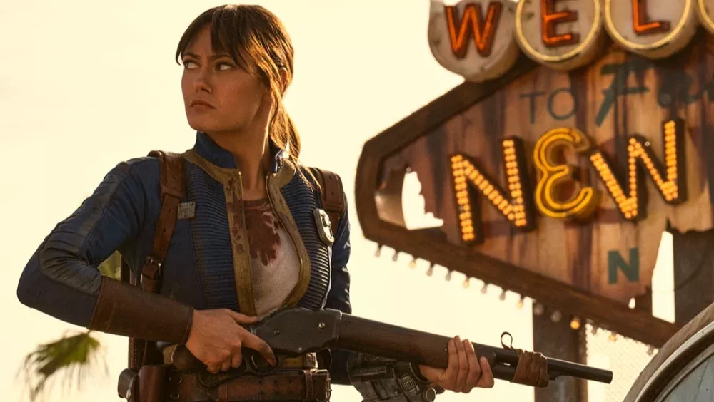 A woman in a blue Vault-Tec jumpsuit holding a shotgun and standing in front of a broken down sign that says "Welcome to Fabulous New Vegas."