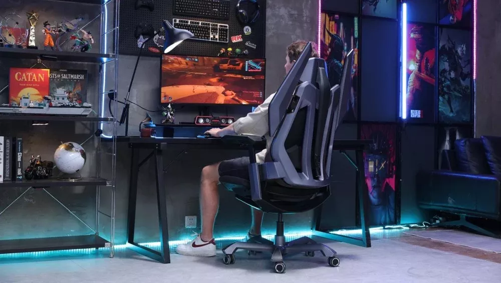 The back of someone sitting in an ergonomic gaming chair. The person is sitting at a desk with a gaming PC and several gaming related items on shelves and the wall near the desk.
