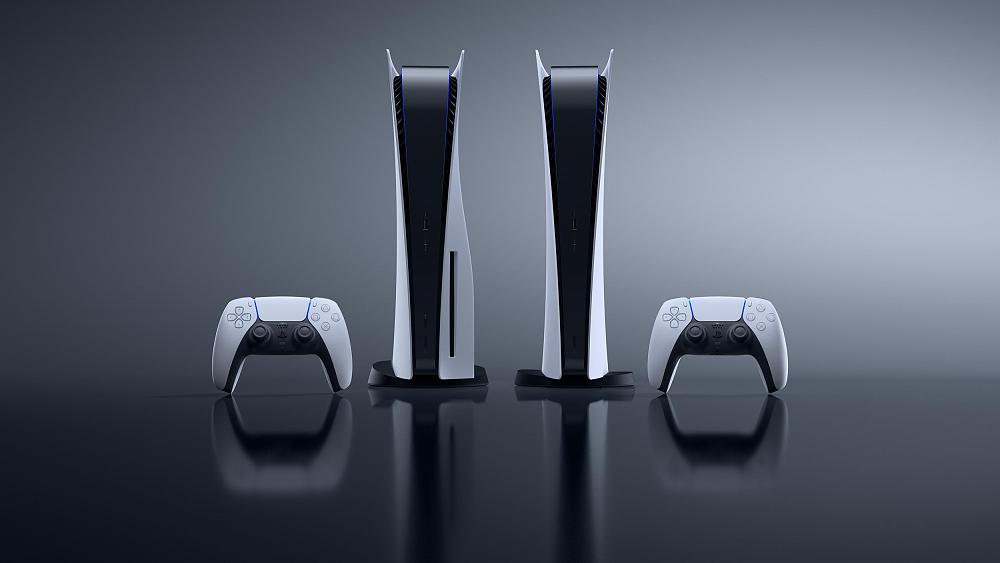 Photo showing two PlayStation 5 consoles and two PlayStation 5 DualSense controllers. The consoles are standing vertically in the center of the image. The controllers are standing upright to the left and right of the consoles.
