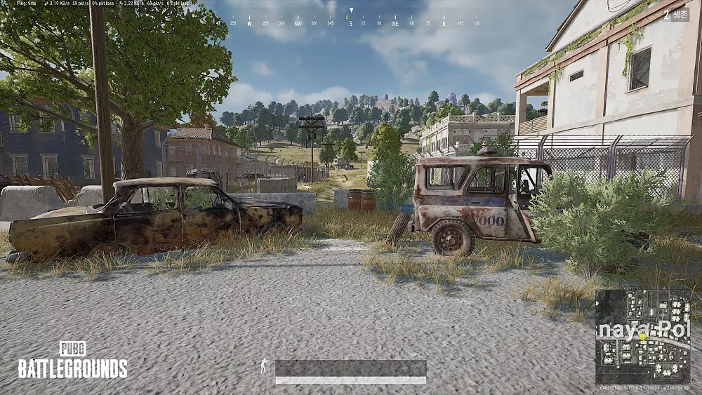 Screenshot from PUBG showing rundown cars, buildings with overgrowth and distant tree covered hills.