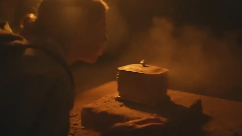 A woman with blonde hair blowing dust off of some sort of ancient artifact in a dimly lit cave.