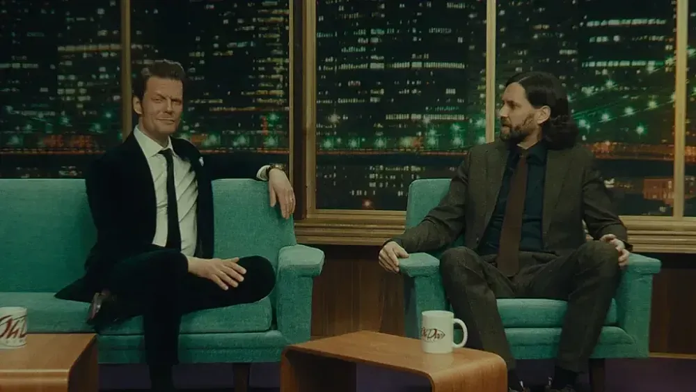 Two men sitting on the set of a talk show, one on the left looking at the camera, the one on the right looking at the man on the left.