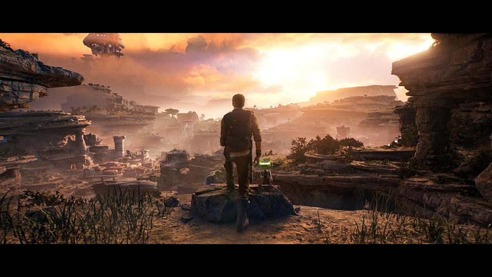 A person stands in a rocky area looking out towards a sunset and a town built near a cliffside in the distance. There is a small robot with green lights right next to the person.