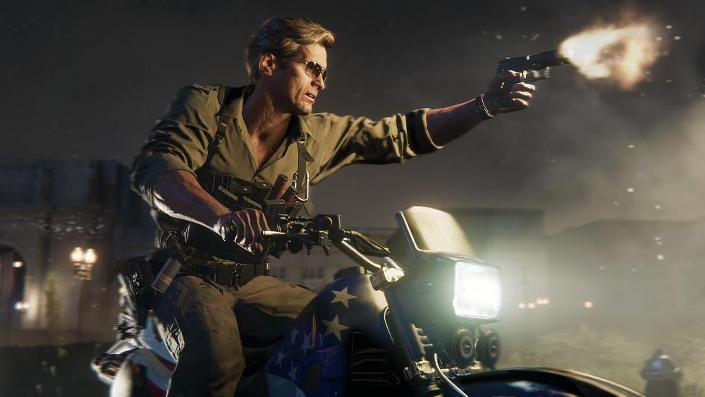 A blonde haired man riding an American flag style motorcycle and firing off a pistol in front of him.