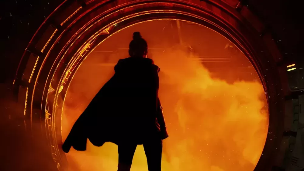 A silhouette of a woman wearing a cape as she stands facing a circular doorway leading to a room full of fire, smoke, and cinder.