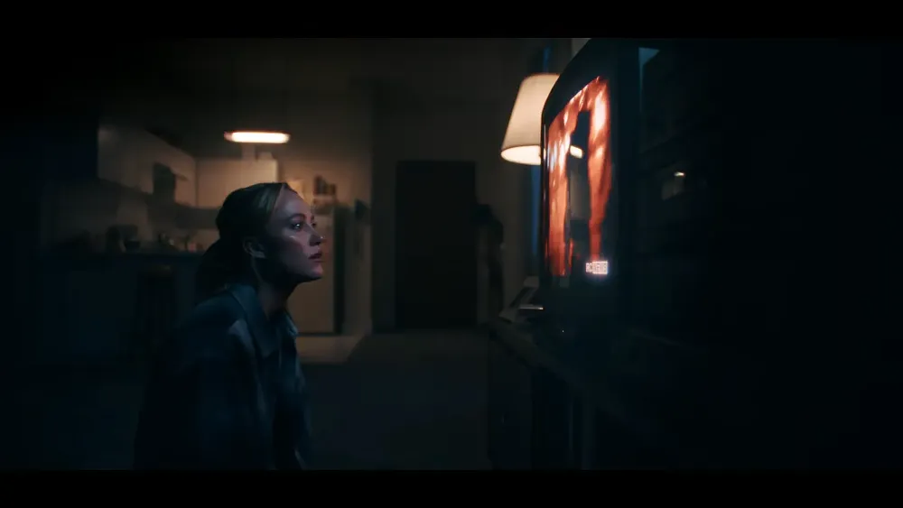 Screenshot from the live-action trailer for Resident Evil Requiem showing a mother sitting on the floor staring at an emergency news broadcast.