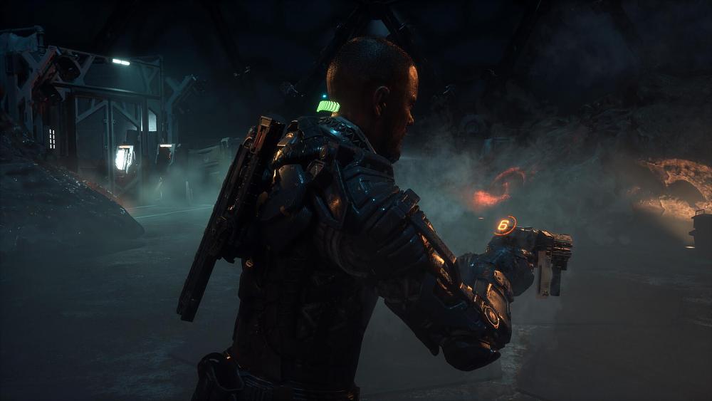 A nearly bald man wearing most of a space suit minus a helmet, readies a pistol-like weapon in his hands while another weapon is attached to his back. He appears to be in some sort of damp, dimly lit cave.
