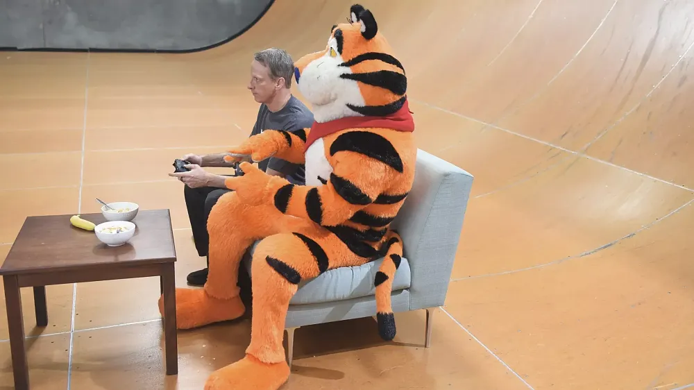 Tony Hawk and Tony the Tiger sitting on a couch on a skateboard vert ramp playing a game together with bowls of cereal sitting on a table in front of them.