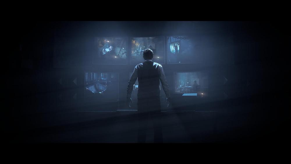 A man with his back to the camera is facing several different security monitors arranged on a wall.