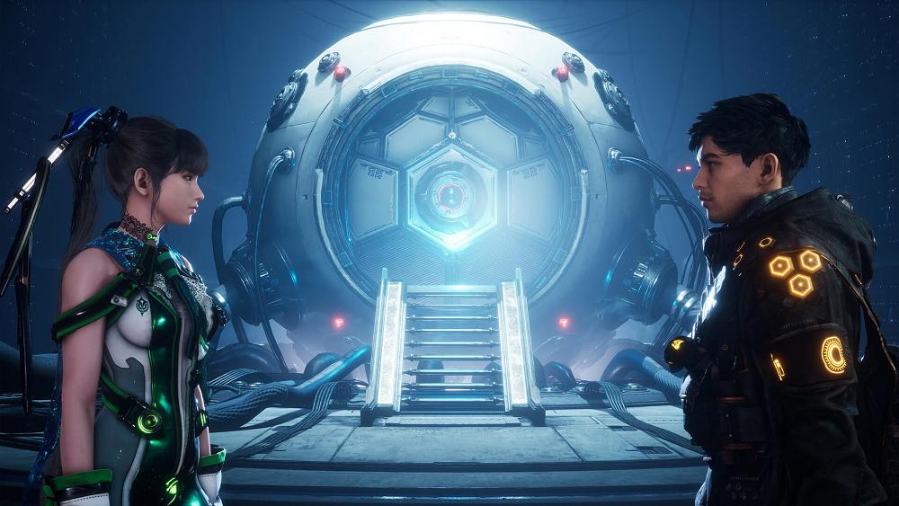 A woman in a green and white skin suit is staring at a man in a black coat with yellow lights. Behind them is some sort of a sphere device connected with a bunch of large wires.
