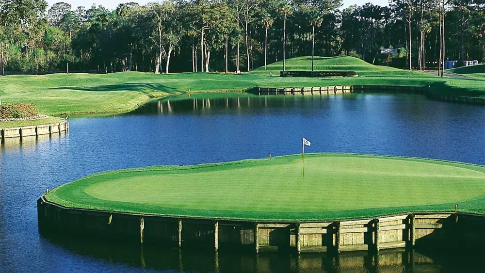 A photo of a golf course showing a green surrounded almost entirely by a lake with trees dotting the background.