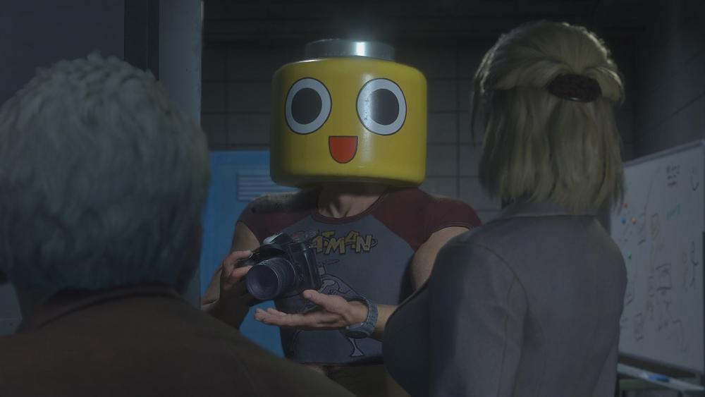 Two people in the foreground looking back towards a man with a very small t-shirt, holding a camera, and wearing a Servbot helmet over his head.