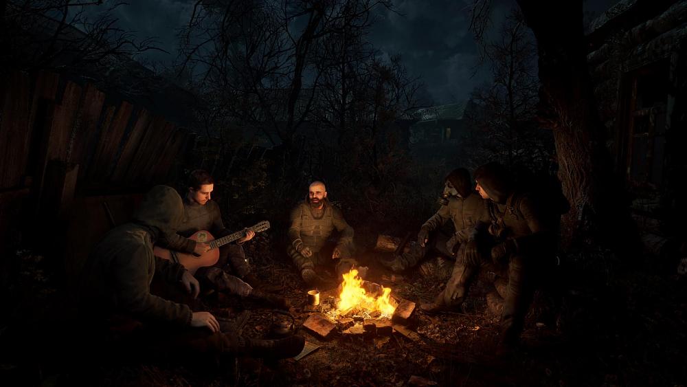 Screenshot showing several men sitting around a camp fire at night near trees. One is playing a guitar. They're all dressed in black or dark, thick clothing. One is wearing a gas mask.