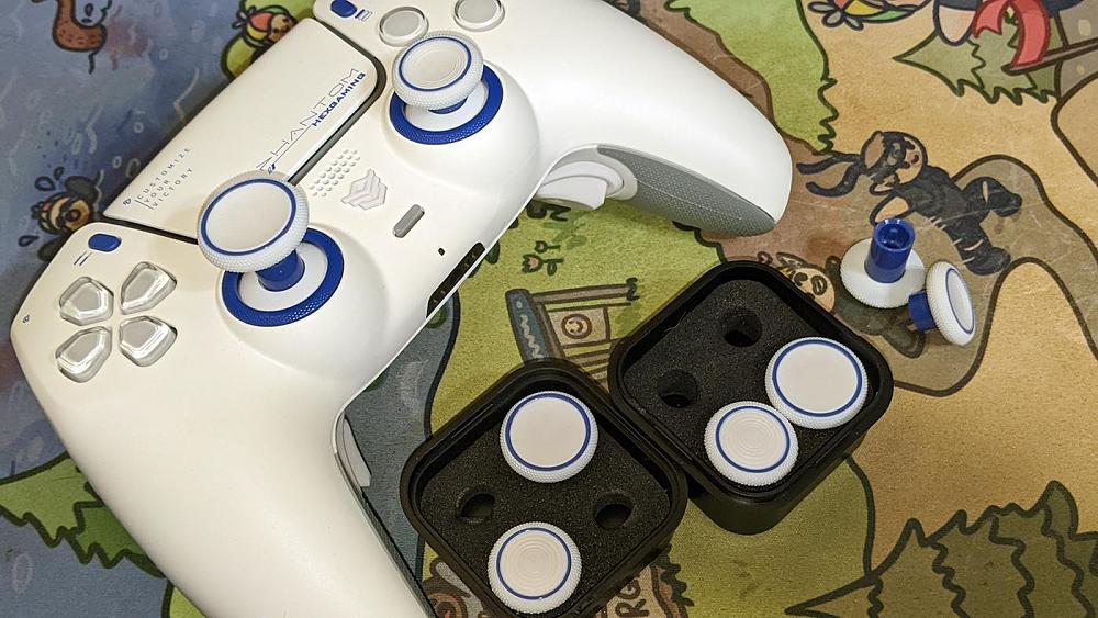 Photo of a custom white with blue accents PlayStation 5 controller from HexGaming, with several swappable thumb sticks next to it.