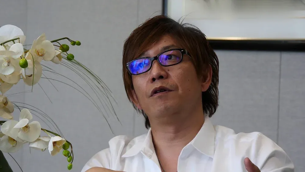 Photograph of a man in the middle of talking. The man is wearing a white shirt. There are some white flowers on the left side of the image.
