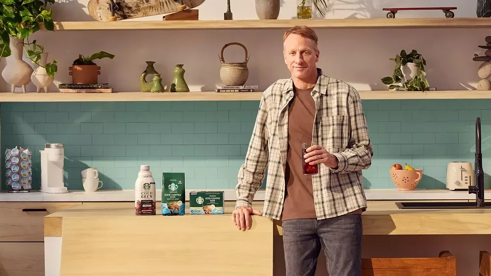 A photograph of a middle-aged Tony Hawk standing in a kitchen with a glass of iced tea.