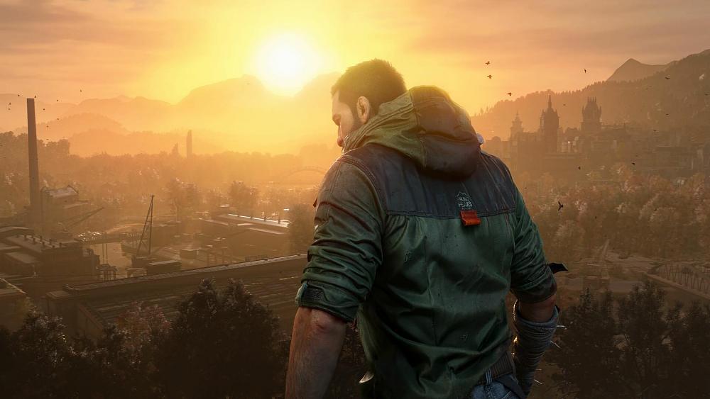 A man in a jacket looking out on a city and forested area as the sun sets in the background behind some hills.
