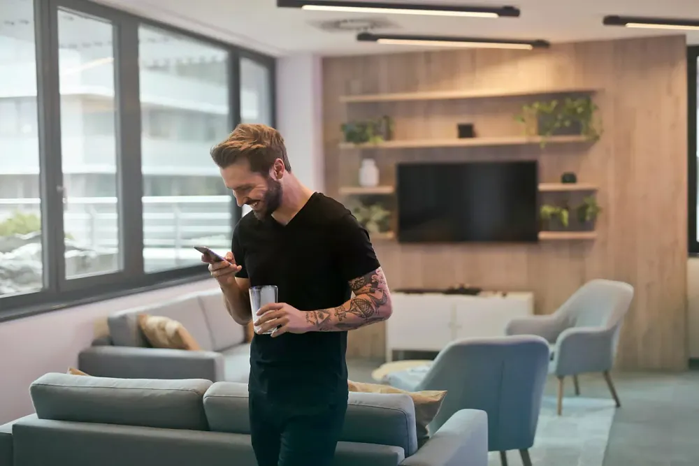 A man with tattoos on his arm looking at his phone in one hand, holding a glass in the other hand, and standing in an apartment.