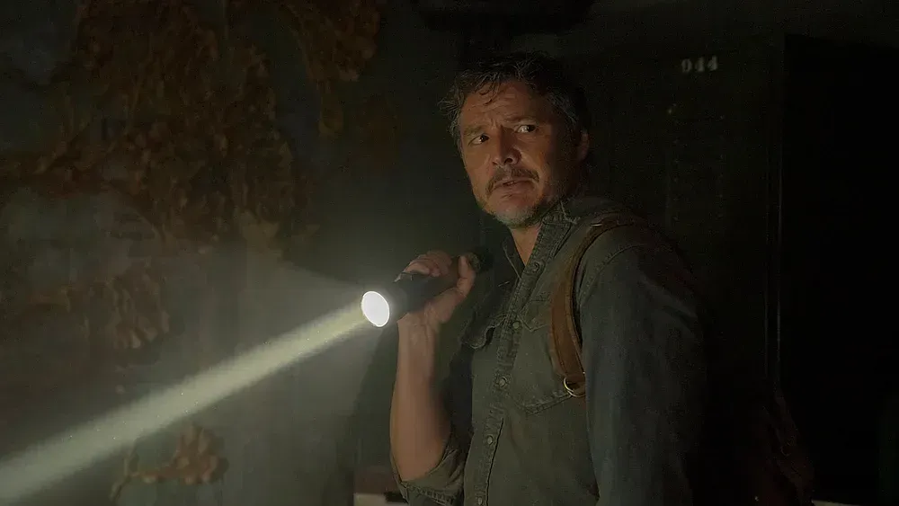 Middle-aged man holding a flashlight in his right hand. He looks off to the side with a worried expression. He is wearing a backpack over a light blue, possibly denim buttoned shirt. He stands near a wall covered in fungi.