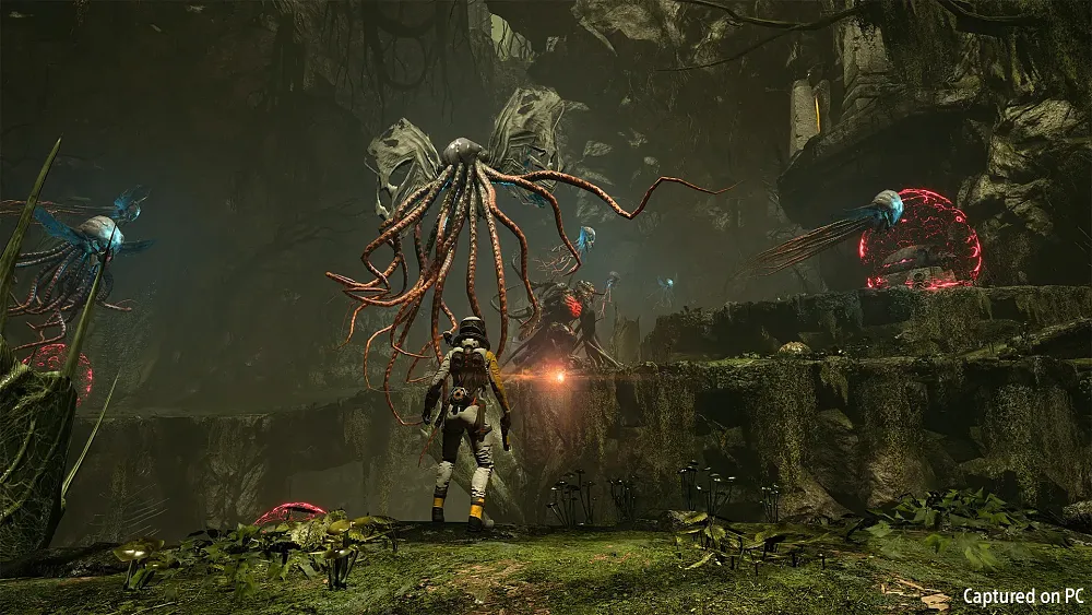 Screenshot from a game, Returnal, running on PC. A character in a space suit stands facing away from the camera. They are holding a gun in their right hand. They are in a green, mossy environment. There are several flying alien creatures flying around, each having about a dozen long tentacles.
