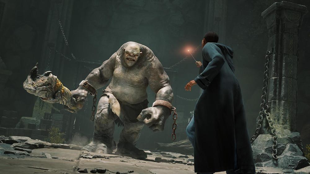 A large troll holding a part of a tree trunk as a club is stomping towards a young wizard in a black robe. The wizard is holding a wand at the ready towards the troll. They look to be fighting in some sort of a dungeon with a couple of stone pillars.