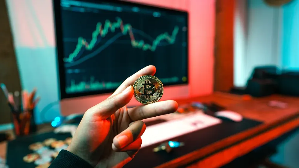 A hand holding a physical Bitcoin in front of a blurred background of a computer and monitor sitting on a desk in a room.