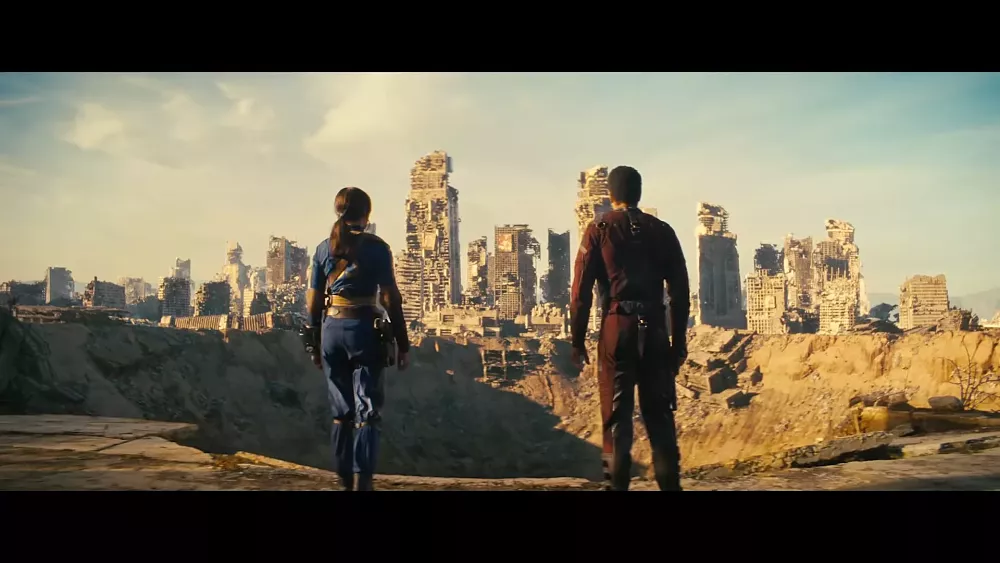 Two people standing near the edge of a massive crater as a ruined city lies in the background.